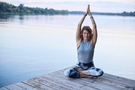 A calm woman in the lotus pose sits on a dock and practices yoga breathing near the river.の写真素材
