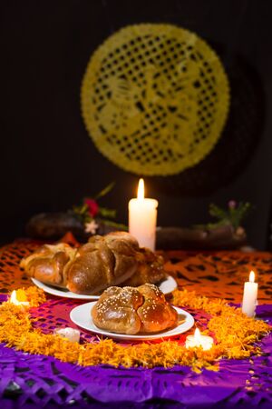 pan de muerto, placed on white plates, adorned with cempasuchil petals and burning candles around...