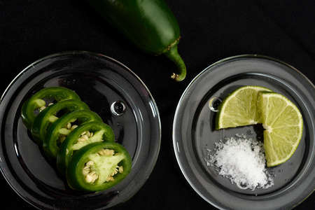 JalapeÃ±o pepper whole and cut into slices, placed in a glass container, around it lemon cut with salt in a glass plate and placed all on a black clothの写真素材