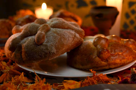 Pan de Muerto, traditional and sugary on a white glass plate, around it marigold flowers, lighted candles, lit copal, diffuse background of colored confetti.の写真素材