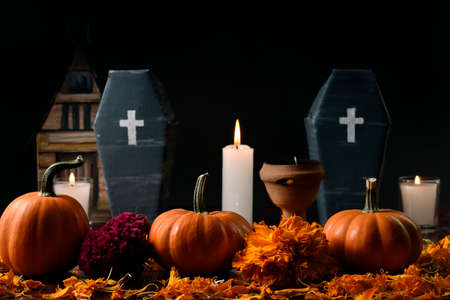 Three pumpkins surrounded with marigold flower, lighted candles and two vertically cardboard coffins and a copal, in the background in diffuse a house made of cardboard.の写真素材