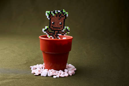 Animated figure of a plant in hama beads, placed in a plastic pot and surrounded by ornamental stones, olive green background.の写真素材
