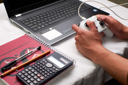 Young man plays a video game with a wired control on his laptop while leaving school aside in times...