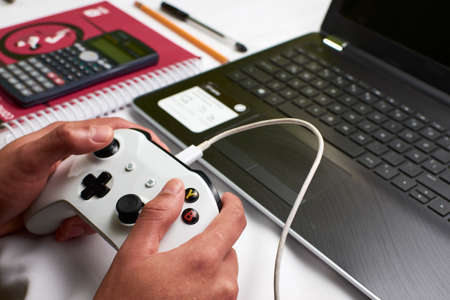 Young man plays a video game with a wired control on his laptop while leaving school aside in times of pandemic, in the background school supplies such as a notebook, calculator, pencil and laptopの写真素材