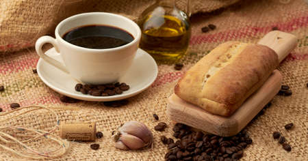 Rustic bread placed on a cutting board, around it a cup of American-style coffee, olive oil, coffee beans and garlic, placed in a vegetable fiber sackの写真素材