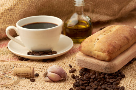 Rustic bread placed on a cutting board, around it a cup of American-style coffee, olive oil, coffee beans and garlic, placed in a vegetable fiber sackの写真素材