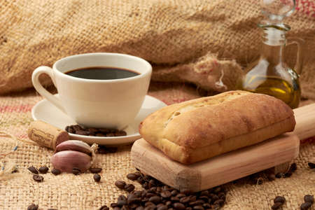 Rustic bread placed on a cutting board, around it a cup of American-style coffee, olive oil, coffee beans and garlic, placed in a vegetable fiber sackの写真素材