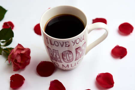 Cup of Americano type coffee, with the legend on the cup "I love you very much mom", adorned with a rose and petals aroundの写真素材