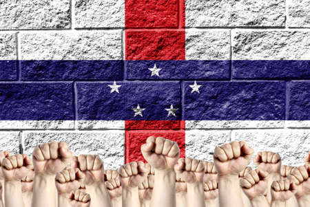 Raised fists against the background of the depicted flag of Netherlands Antilles on a brick wall, a concept of strength and unity.の写真素材