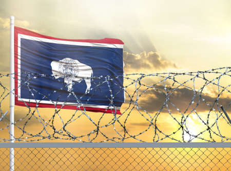 Flagpole with the flag of State of Wyoming against the sky and behind a fence with barbed wire. The concept of protecting the borders of territories.の写真素材