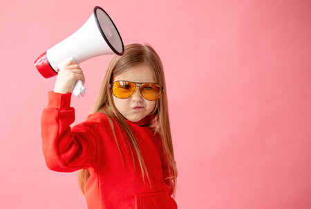 Little girl in red jacket and sunglasses with megaphone on pink backgroundの写真素材