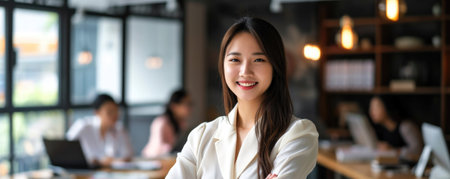 An asian woman stands confidently in an office space with large windows. She is smiling, and there is a busy work atmosphere in the background with colleaguesの素材
