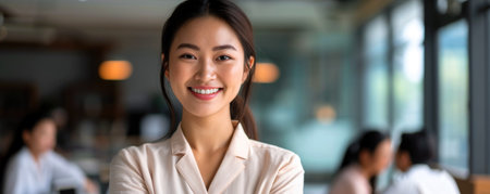 An asian woman stands confidently in an office space with large windows. She is smiling, and there is a busy work atmosphere in the background with colleaguesの素材