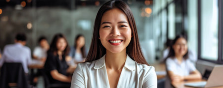 An asian woman stands confidently in an office space with large windows. She is smiling, and there is a busy work atmosphere in the background with colleaguesの素材