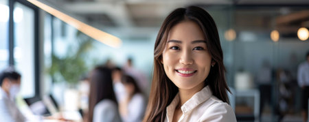 An asian woman stands confidently in an office space with large windows. She is smiling, and there is a busy work atmosphere in the background with colleaguesの素材