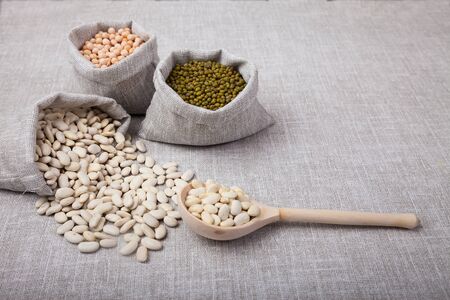 white beans spilled from the bag, mung bean and peas in the bag and a wooden spoon lie on a linen backgroundの写真素材
