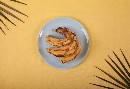 Ugly bananas lie on a blue porcelain plate on a yellow background with palm leaves .の写真素材