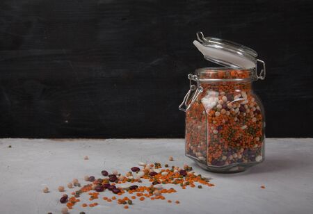 White and red beans, peas, green and red lentils in an open glass jar and scattered legumes on on a...