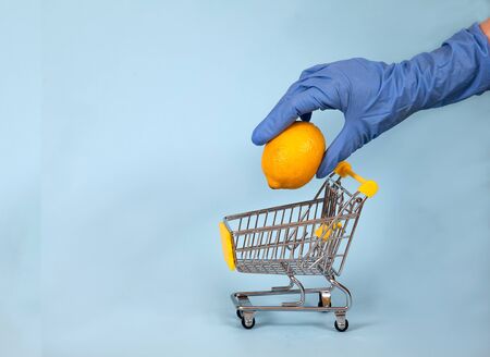 Female hand in a blue rubber glove lowers a lemon into a supermarket trolley on a blue background. Protection. Hygiene. Health. Copy spaes. covid 19の写真素材