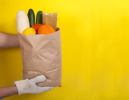 Male hands in cotton gloves hold a paper bag with pasta, tomatoes, cucumbers, daikon, lemon, orange on a yellow background. Zero waste. Courier delivery. Volunteerの写真素材