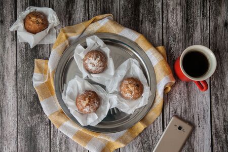 Muffins on a metal plate and coffee in a red ceramic mug, next to a smartphone stand on a wooden...