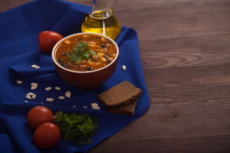 Soup with vegetables and beans, tomatoes, oil in a bottle and rye bread on a blue cloth on a wooden background. Healthy eating.の写真素材