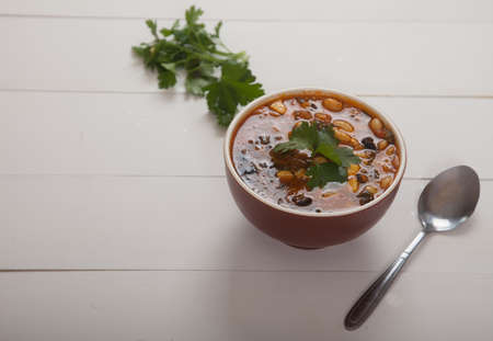 Bean soup with vegetables, olives and cheese, fresh parsley, a metal spoon stand on a white wooden...
