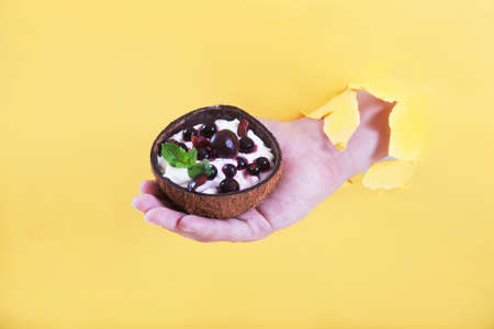 A man's hand in a hole holds a cup of coconut with a dessert of whipped cream and currants and...