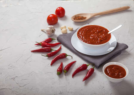 Spicy harissa sauce in a white porcelain cup, chili peppers, garlic and tomatoes on a white concrete background. Copy spaesの写真素材