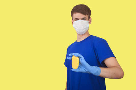Young male courier in a medical mask and rubber gloves holding a box on a yellow background. Social...