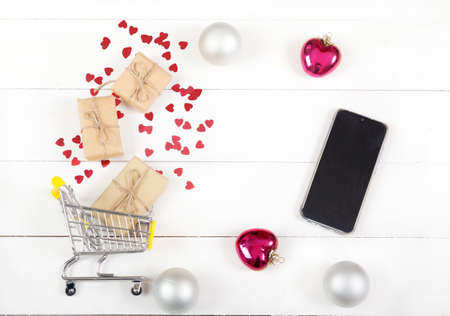 Christmas shopping for gifts by smartphone. Grocery cart with gifts, balls, hearts and blank phone on a white wooden background. Ordering gifts on the Internet. online shoppingの写真素材