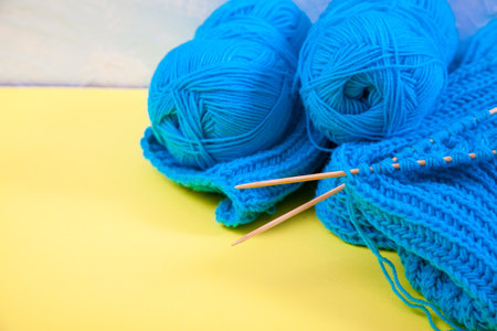A rolled up hand-knitted blue scarf, balls of yarn and knitting needles lie on a yellow backgroundの写真素材