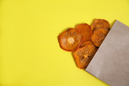 Persimmon chips in a bag of craft paper on a yellow background. Healthy food conceptの写真素材