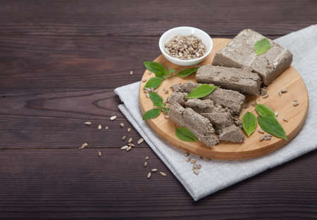 Briquettes of sunflower halva, sunflower seeds and green leaves lie on a wooden board on a gray...
