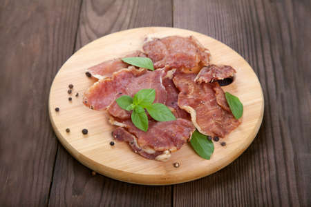 Pieces of jerky meat with basil leaves lie on a wooden cutting board on a wooden background.の写真素材