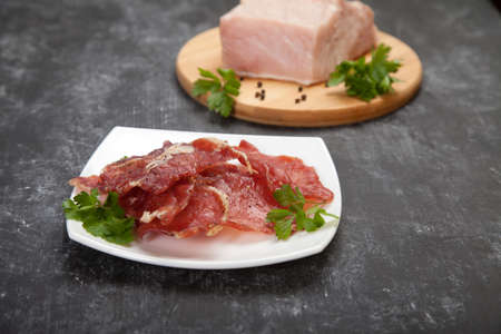 Cured meat on a white plate and raw meat with herbs on a wooden cutting board on a black background.の写真素材