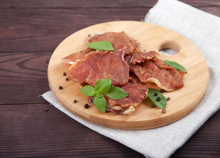 Pieces of jerky with basil leaves lie on a wooden cutting board, under it is a linen napkin on a wooden background.の写真素材