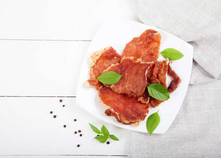 Pieces of jerky, basil leaves, black peppercorns lie on a white plate on a white wooden background. Copy spaes.の写真素材