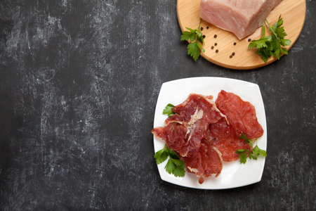 Pieces of jerky on a white porcelain plate and raw meat on a cutting board on a black background. Copy spaes.の写真素材