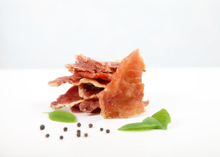 Pieces of jerky, basil leaves and black pepper on a white plate.の写真素材