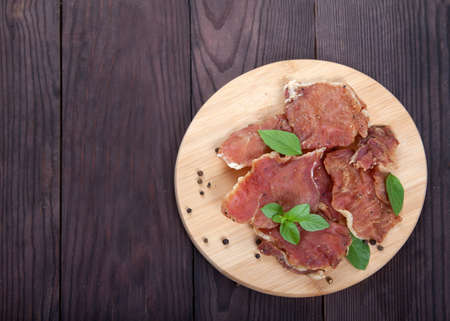 Pieces of jerky meat with basil leaves lie on a wooden cutting board on a wooden background.の写真素材