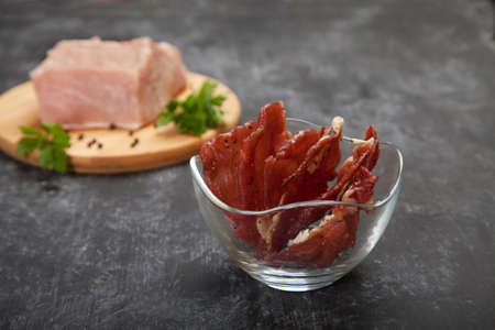 Pieces of jerky in a glass cup and raw meat on a cutting board on a black background.の写真素材