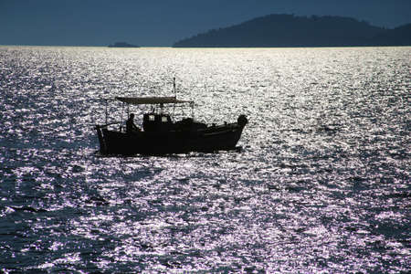 A fishing boat on the glistening open sea. In the distance the mountains of the island. Sunset.の写真素材