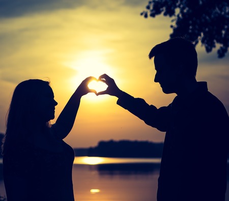 Couple doing heart shape with their hands on lake shore. People silhouettes vintage photo.の写真素材