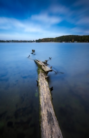 Long exposure landscape of lake shore with dead tree trunk and driftwood. Lake Krzywe in Olsztyn, Poland.の写真素材