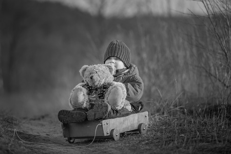 Young happy boy playing with his teddy bear and carriage toy outdoor in beautiful rural landscape in golden light at spring. Happy childhood spent in the countryside.の写真素材