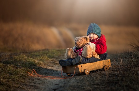 Young happy boy playing with his teddy bear and carriage toy outdoor in beautiful rural landscape in golden light at spring. Happy childhood spent in the countryside.の写真素材