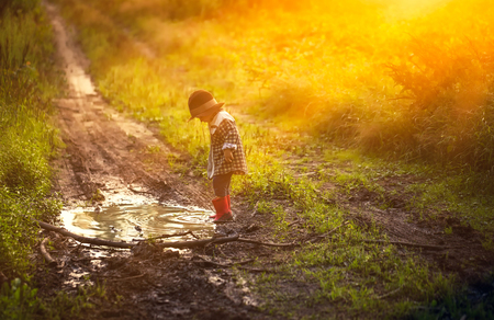 Little boy in hat and rubber shoes playing in puddle in summer forest.の写真素材