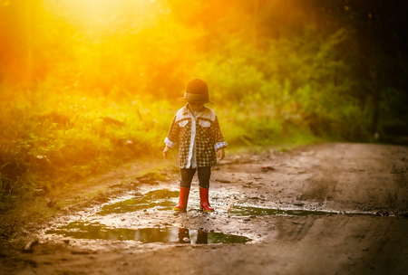 Little boy in hat and rubber shoes playing in puddle in summer forest.の写真素材