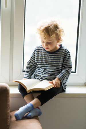 Young caucasian boy with curly hair reading book and sitting on windowsill.の写真素材